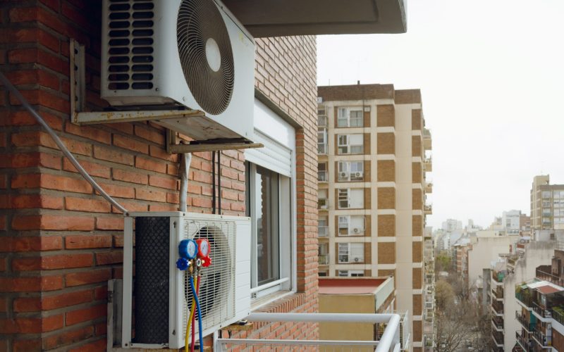 Two air conditioning units being maintained on a brick wall installed on the balcony of an apartment, there are residential buildings in the background with the sky and the city of Buenos Aires.